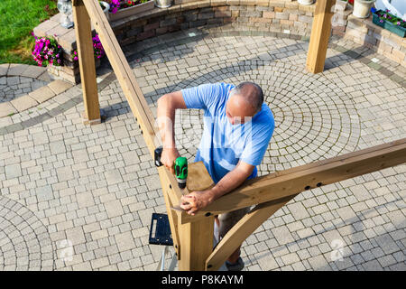 Man building a wooden gazebo on a brick patio with ornamental paving viewed from above as he stands on a ladder screwing together beams Stock Photo