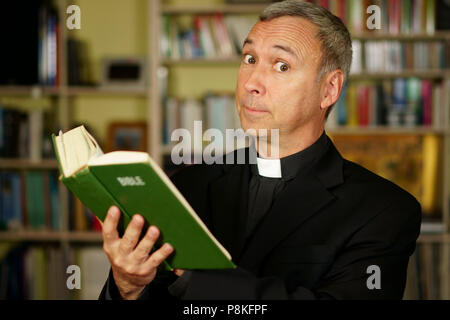 A good looking serious catholic priest is studying, reading the Bible, into his library. He looks at us with desapprovation. Stock Photo