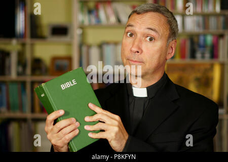 A good looking serious catholic priest is studying, reading the Bible, into his library. He looks at us with disapproval. Stock Photo