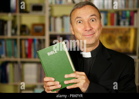 A good looking serious and crazy catholic priest is studying, reading the Bible, into his library. He looks at us with a foolish approbation. Stock Photo