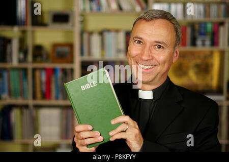 A good looking catholic priest is studying, reading the Bible, into his library. He looks at us with serenity and optimism. Stock Photo