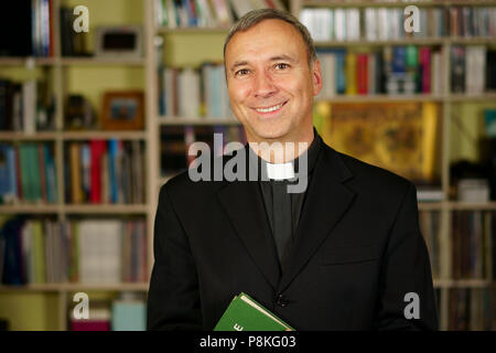 A good looking catholic priest is studying into his library. He looks at us with serenity and optimism. Stock Photo