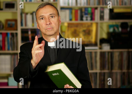 A good looking catholic priest is giving the blessing into his library. He seriously looks at us with  interest, disapproval, judgement, pensiveness a Stock Photo