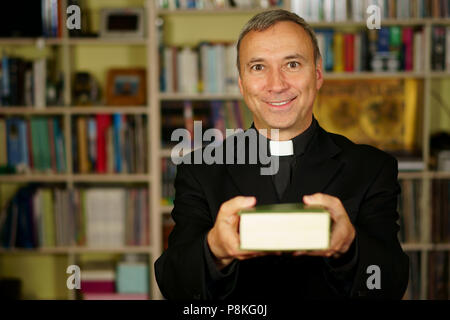 A good looking catholic priest is studying into his library. He looks at us with serenity, optimism and is offering the Bible. Stock Photo