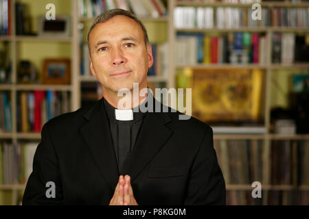 A good looking catholic priest is praying into his library. He looks at us with serenity, optimism, joy, admiration, trust, confident and peace. Stock Photo