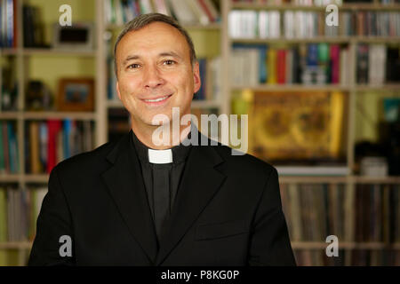 A good looking catholic priest is studying into his library. He looks at us with serenity, peace and optimism. Stock Photo