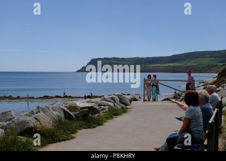 View across the sea to Ravenscar cliffs from Robin Hood's Bay,Yorkshire Heritage Coast,England,UK with people seated on bench and walking to sea Stock Photo