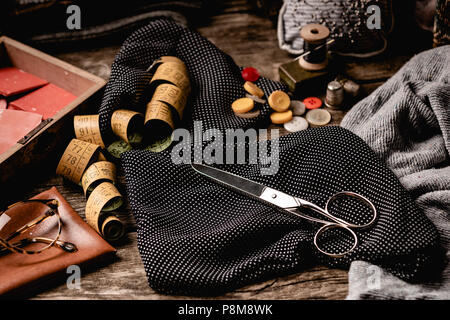 Composition of black polka-dotted fabric with scissors, sewing spools, tape measure and colorful buttons with glasses placed on rough wooden table Stock Photo