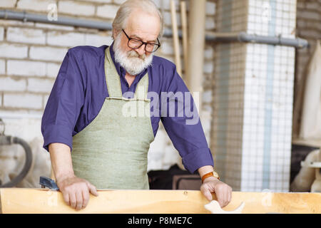 close up portrait of old craftsperson in glasses with shirt and apron holding plank to lay it on the floor of the garage Stock Photo