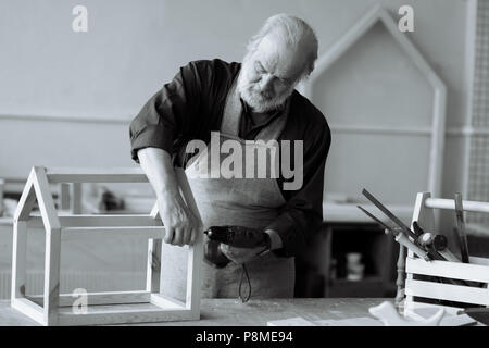 white and black photo of old grey haired carpenter building feeder for birds in the repair shop Stock Photo
