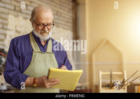attractive old carpenter working on designs in his workshop.close up photo. write down the plan of work Stock Photo