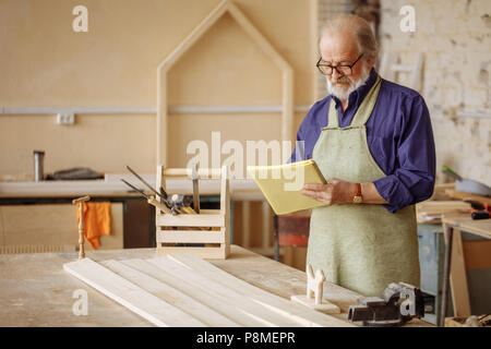 old grey headed joiner in work clothes is concentrated on making up project. making up plan. copyspace Stock Photo