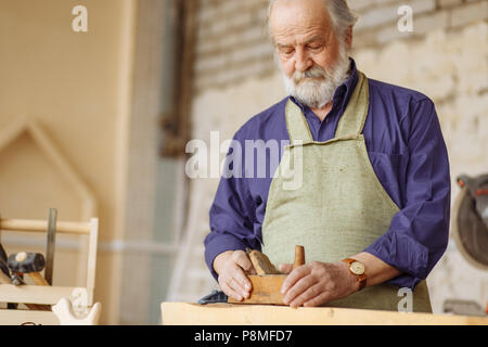 old experienced carpenter working with wooden planer on plank in workshop. Stock Photo