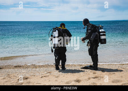 Two deep sea divers getting ready to dive by performing safety checks on their equipment together on a beach with the ocean behind Stock Photo