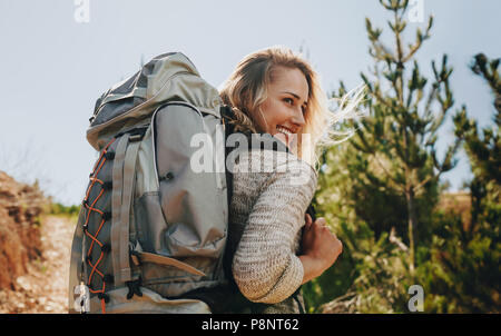 Rear view shot of smiling woman with backpack going on a camping. Caucasian female hiker on mountain looking away and smiling. Stock Photo