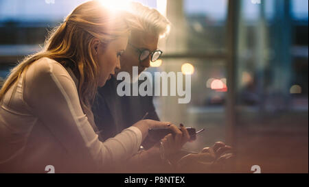 Business colleagues sitting together in office lobby and using their mobile phones. Business professionals using cell phones during a break in office. Stock Photo