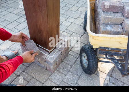 Man building a brick pillar on a wooden gazebo leg carefully placing one in position after applying the glue Stock Photo