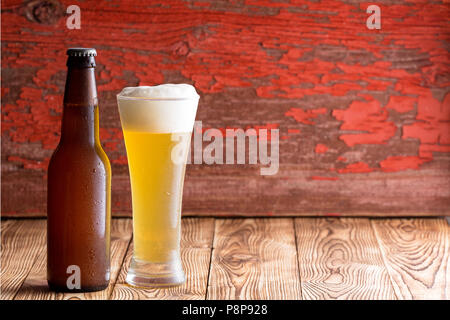 Ice cold frothy pale ale in a tall glass alongside an unlabelled beer bottle with long neck over a rustic wood background with peeling red paint and c Stock Photo