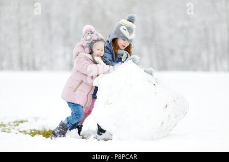 Two funny adorable little sisters making a snowman together in beautiful winter park during snowfall Stock Photo