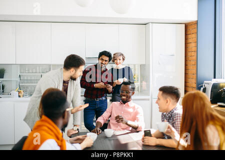 cheerful young Indian man and elegant Caucasian woman standing and looking at colleagues who are sitting at the table Stock Photo