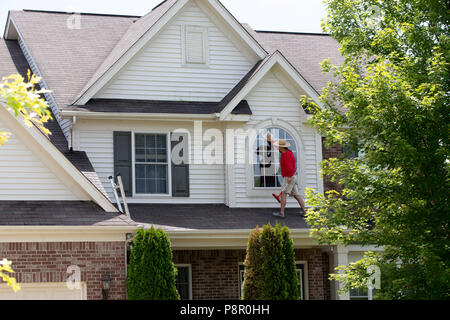 Window cleaner cleaning a large arched view window on the second floor facade of a house standing on the roof of the patio below Stock Photo