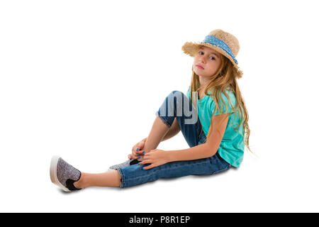 Solemn thoughtful little girl with long blond hair wearing a trendy straw hat sitting on the ground in a relaxed position watching the camera isolated Stock Photo