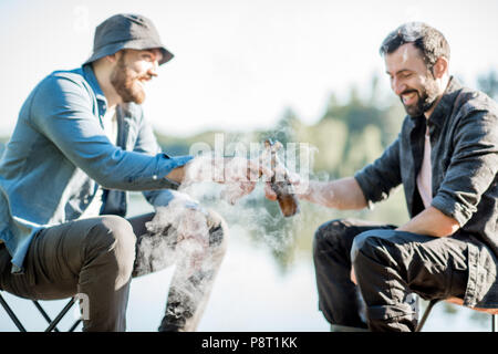 Two fishermen frying fish sitting with beer during the picnic on the wooden pier near the lake in the morning Stock Photo