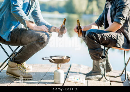 Two fishermen frying fish sitting with beer during the picnic on the wooden pier near the lake in the morning Stock Photo