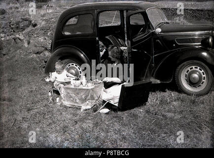 1950s, historical, a day out ....mother with young child sitting by their motor car having a civilised picnic set up on a small table with tablecloth, England, UK. Stock Photo
