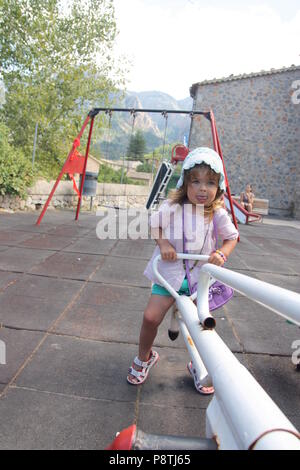 Beautiful young toddler girl in a bonnet and sandals on a seesaw in a park concentrating on balancing with her tongue sticking out Stock Photo