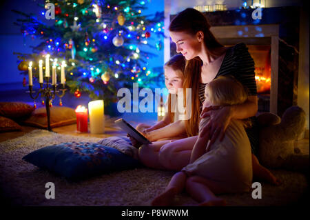 Young mother and her daughters using a tablet pc by a fireplace on warm Christmas evening Stock Photo