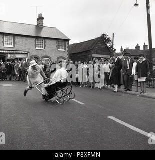 1967, historical, people watching a pram race - using traditional prams or baby carriages - taking place on a road at a village fair at Quainton, Buckinghamshire, England, UK. Stock Photo