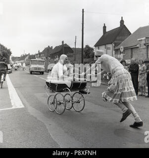 1967, historical, people watching a pram race - using traditional prams or baby carriages - taking place on a road at a village fair at Quainton, Buckinghamshire, England, UK. Stock Photo