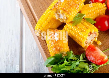 Tasty Barbecued corn cobs with herbs, vegetables and salt on a board and a white wooden table Stock Photo