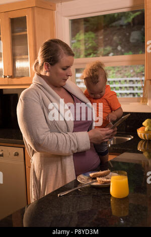Mother with baby standing in the kitchen and using digital tablet Stock Photo