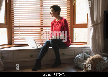 Woman looking through window while using laptop in living room Stock Photo
