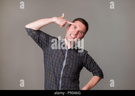 Tired or bored european guy holding gun gesture near temples and making faces while standing over gray background. Stock Photo