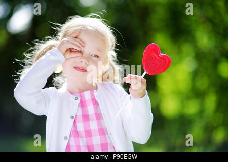 Cute little girl eating huge heart-shaped lollipop outdoors on beautiful summer day Stock Photo