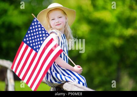 Adorable little girl wearing hat holding american flag outdoors on beautiful summer day. Independence Day concept. Stock Photo