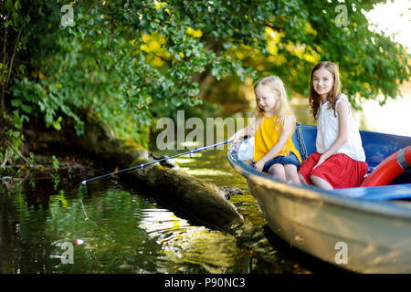 Two cute little girls having fun in a boat by a river at beautiful summer evening. Sisters fishing with a fishing rod on warm and sunny day. Stock Photo