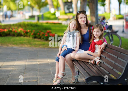 Young mother and two cute little sisters sitting on a bench hugging on warm and sunny summer day in italian town Stock Photo