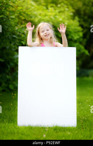 Cute little girl holding big blank whiteboard on warm and sunny summer day outdoors Stock Photo