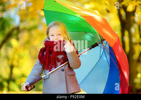 Cute little girl holding rainbow umbrella on beautiful autumn day. Happy child playing in autumn park. Kid gathering yellow fall foliage. Autumn activ Stock Photo