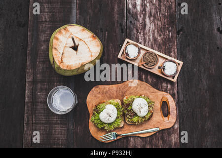 Tasty organic healthy breakfast - poached eggs on toast with avocado, water with lime, young coconut. Dark wooden background Stock Photo