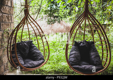 Two rattan hanging wicker chairs with black pillows in garden, summertime Stock Photo