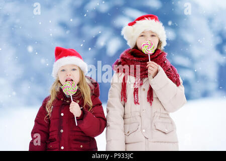Two adorable little sisters wearing Santa hats having huge striped Christmas lollipops on beautiful winter day. Children with lolly candies on Xmas da Stock Photo