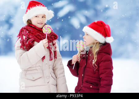 Two adorable little sisters wearing Santa hats having huge striped Christmas lollipops on beautiful winter day. Children with lolly candies on Xmas da Stock Photo