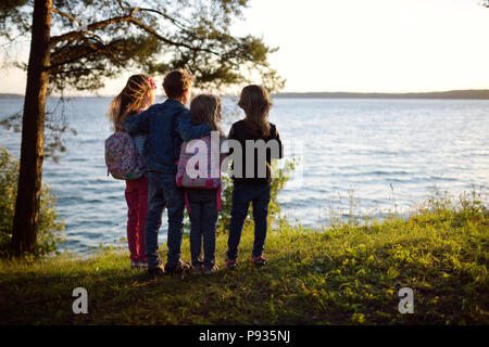 Four cute little sibling standing by the lake enjoying beautiful sunset view. Children exploring nature. Family activities in summer. Stock Photo