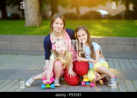 Cute portrait of young mother and two children riding skateboards in a city park on sunny summer evening. Active leisure and outdoor sport for kids. Stock Photo