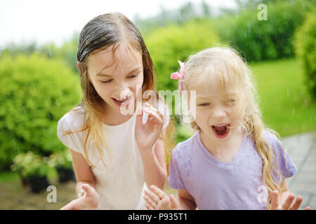 Two cute little sisters having fun under warm summer rain. Siblings playing together outdoors. Stock Photo